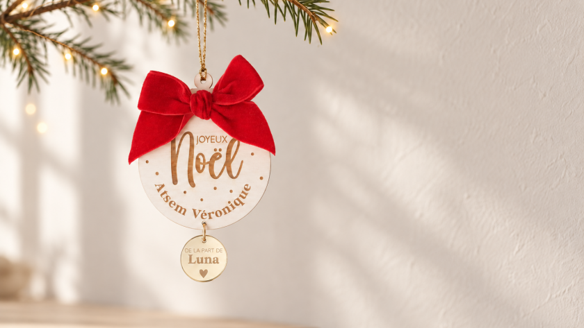 Boule de Noël personnalisée en bois avec nœud rouge et médaillon doré, suspendue à une branche de sapin illuminée