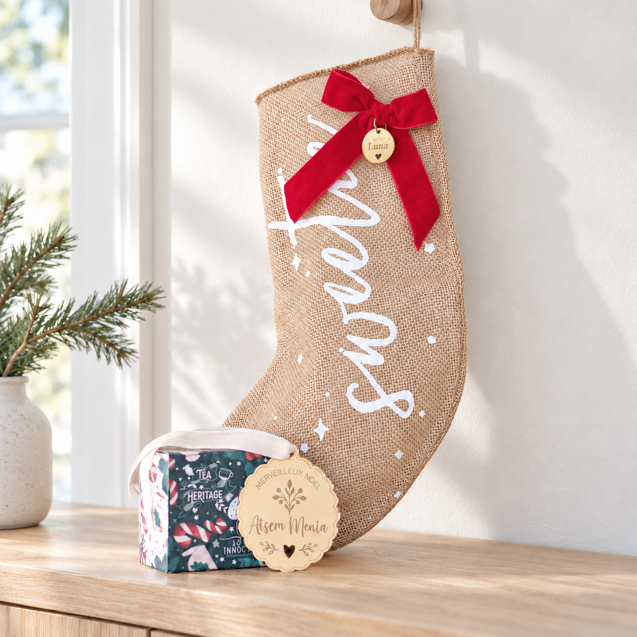 Chaussette Sweetie en jute à nœud rouge, coffret de thé et boule personnalisée dorée près d’une fenêtre